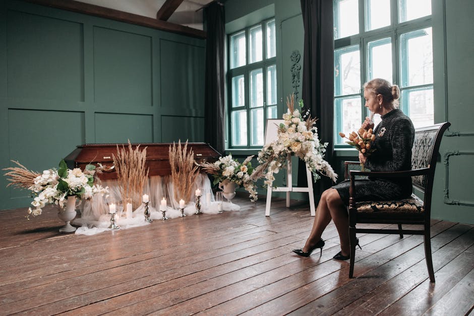 A woman sits on a floral-patterned upholstered wooden chair in a softly lit room with green paneled walls and large windows. She is dressed in a dark, textured dress with high heels and holds a bouque