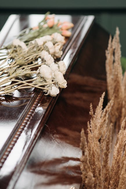 A delicate floral arrangement resting on a polished wooden surface, consisting of small, white, dried flowers with slender stems, accented by soft pink tulips with closed buds and green leaves. The bo