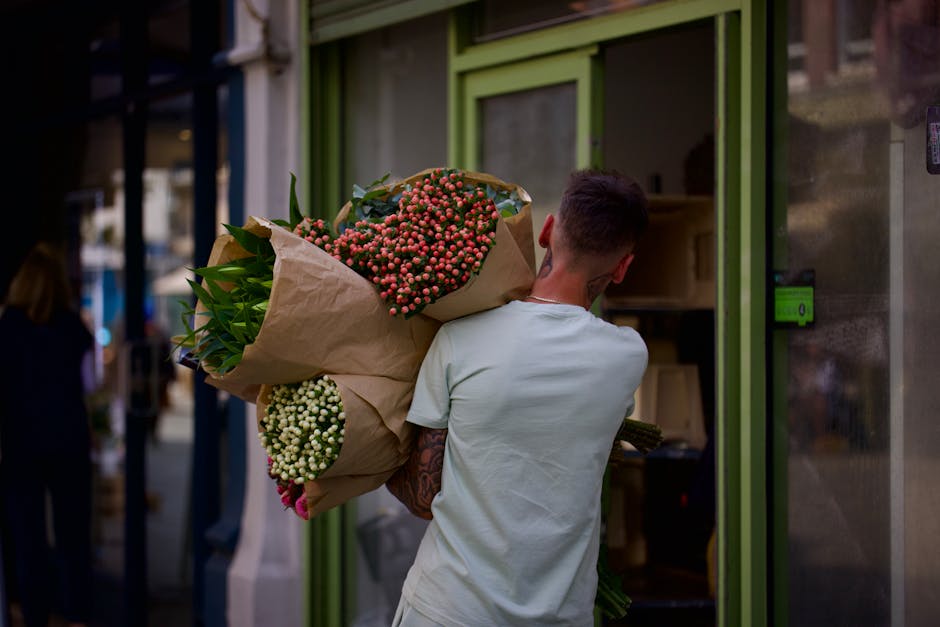 A person with short dark hair, wearing a light grey t-shirt, is seen from the back carrying a large bouquet of fresh flowers near a green storefront. The bouquet includes pink and white clustered flow