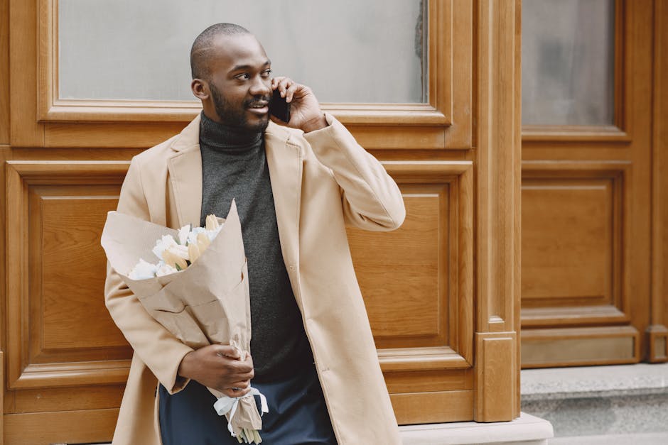 A man with dark skin and a neatly trimmed beard is standing outdoors in front of a wooden-panelled building, holding a bouquet of fresh white flowers wrapped in beige paper with a white ribbon. He is 