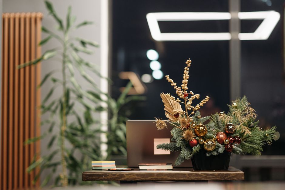 A festive floral arrangement featuring a mix of evergreen branches, gold and bronze baubles, dried flowers, and berry accents, arranged in a black vase placed on a wooden table. The bouquet includes s