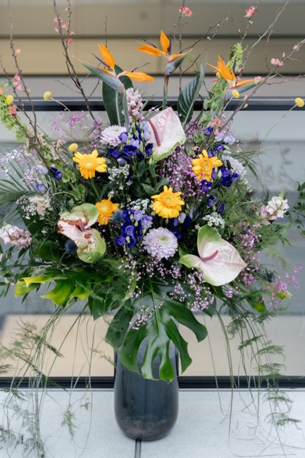 A tall, lush floral arrangement in a dark, cylindrical vase featuring a mix of fresh flowers including large white calla lilies with pink-tinted edges, vibrant yellow daisies, deep blue asters, and de