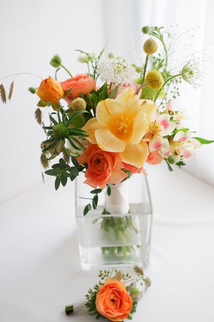 A vibrant floral arrangement in a clear glass vase, featuring large yellow amaryllis, soft peach roses, pink and white alstroemeria, and various greenery and small round flower accents. The bouquet is