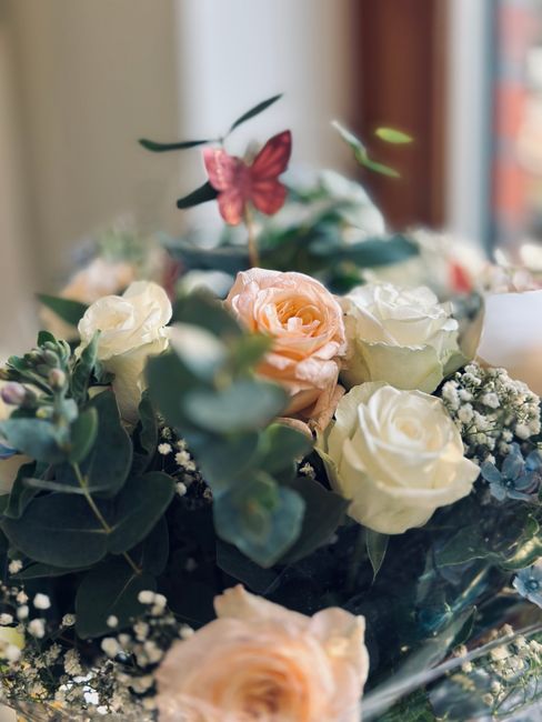 A close-up of a floral arrangement featuring soft peach roses, white roses, and sprigs of baby's breath, accented with dark green eucalyptus leaves. The flowers are arranged in a glass vase, with some